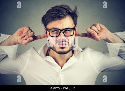 Giovane uomo orinato accigliata e guardando lontano durante la chiusura di orecchie irritazione su sfondo grigio Foto Stock