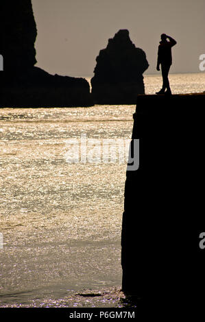 Un uomo in piedi su un Cornish seawall al tramonto guardando il mare stagliano contro il cielo Foto Stock