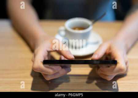 Giovane donna utilizzando un telefono cellulare come ella beve caffè presso un ristorante tavola in una vista ravvicinata sulla parte superiore del dispositivo Foto Stock