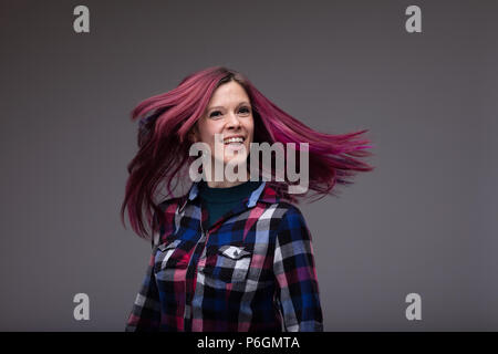 Spensierato ridere la donna in un elegante plaid top sfogliare i suoi lunghi tinti capelli viola come ella ghirigori intorno isolato su grigio Foto Stock