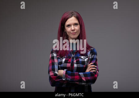 Considerato attraente giovane donna con un sorriso amichevole e long purple capelli oscurata la sua corrispondenza plaid top in piedi con le braccia incrociate guardando la ca Foto Stock