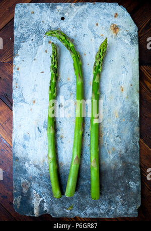 Tre steli di verde di asparagi freschi su un grigio rustico in pietra rettangolare sulla scheda ano vecchio tavolo in legno top Foto Stock