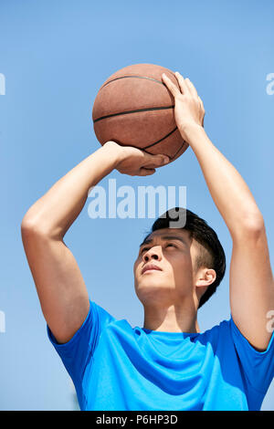 Giovane maschio asiatici giocatore di basket facendo un jump shot contro il cielo blu sullo sfondo. Foto Stock