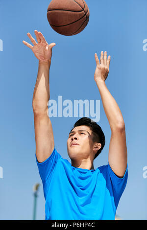 Giovane maschio asiatici giocatore di basket facendo un jump shot contro il cielo blu sullo sfondo. Foto Stock