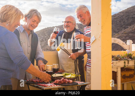 Quattro active senior pensionati hanno divertimento nella terrazza a casa la cottura di alcuni barbecue sorrisi a tutti e di stare insieme in amicizia sotto una bella giornata di sole. Foto Stock