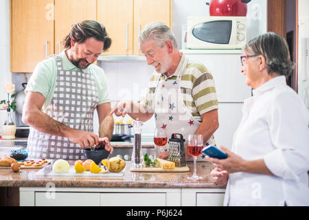 Famiglia caucasica uomo tre persone cucinare insieme lokking il come fare sul telefono cellulare della madre. Tutti divertirsi e ridere preparazione someth Foto Stock