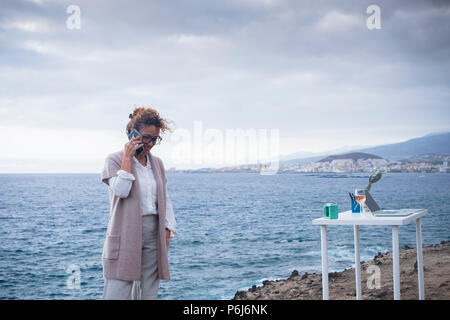 Nuovo stile di vita alternativo opera in naturale ufficio esterno vicino all'oceano. bello stile di vita il sogno di tutti. donna parlare e lavorare con lo smartphone in sc Foto Stock