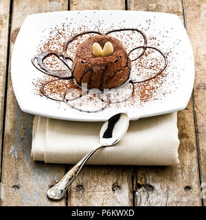 Dolce al cioccolato torta decorata con lo sciroppo al cioccolato mandorle e cacao in polvere sulla piastra bianca accanto a silver piccolo cucchiaio di legno sul tavolo rustico Foto Stock