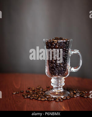 I chicchi di caffè in un vetro trasparente su un tavolo di legno con uno sfondo grigio. Foto Stock