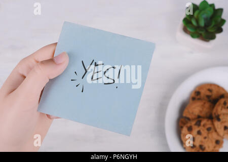 Donna mani tenendo nota di carta con il testo "Sì!" e cookie. Focus su carta Foto Stock