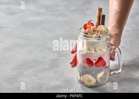 Un sano dolce fatto da organici semi di Chia con yogurt, panna, e fette di banana e le fragole in un bicchiere. Femmina prende mano un bicchiere su un g Foto Stock