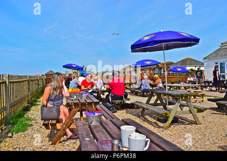 Le persone in un momento di relax a banchi fuori del lungomare café in una calda giornata estiva con un gabbiano flying overhead. Legno tavoli da picnic con ombrelloni aperti. Foto Stock