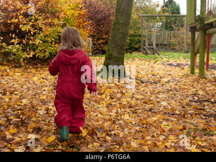 Vista posteriore del bambino a piedi attraverso le foglie di autunno indossando una pozza rosso tuta e wellies verde Foto Stock