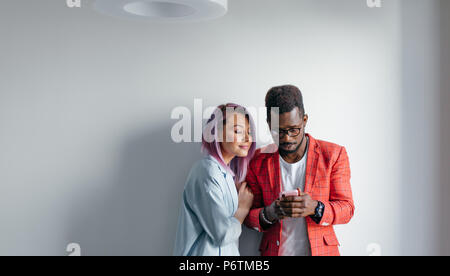 Multirazziale amici la condivisione di foto con ciascun altro, isolato in piedi oltre il muro grigio sfondo, copia dello spazio. Foto Stock