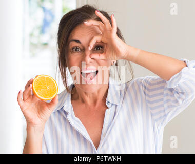 Donna di mezza età tenendo la frutta di arancia con buon viso sorridente facendo segno OK con la mano sull'occhio guardando attraverso le dita Foto Stock