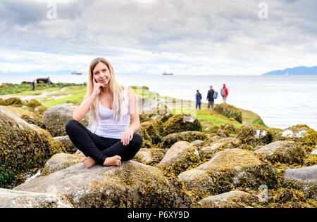 Giovane donna seduta su roccia e sorridente cordiale nella fotocamera in zona intercotidale di Vancouver, British Columbia. Queste rocce sono sott'acqua di Foto Stock