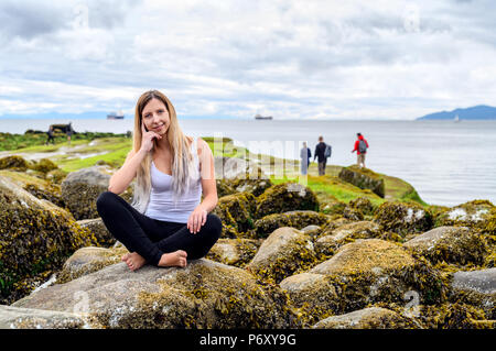 Giovane donna seduta su roccia e sorridente cordiale nella fotocamera in zona intercotidale di Vancouver, British Columbia. Queste rocce sono sott'acqua di Foto Stock