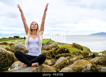 Giovane donna seduta su roccia e le sue braccia sono sollevati in aria in zona intercotidale di Vancouver, British Columbia. Queste rocce sono sott'acqua di Foto Stock