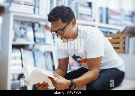 Giovani spectacled brunette uomo seduto e libro di lettura con coperchio bianco Foto Stock