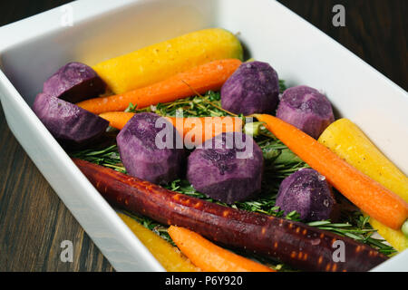 Fresh giallo, viola e arancione carote e patate viola sul cuscino di erbe in una teglia per arrosti Foto Stock