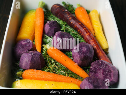 Fresh giallo, viola e arancione carote e patate viola sul cuscino di erbe in una teglia per arrosti Foto Stock