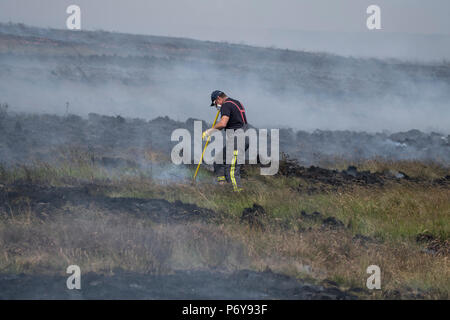 1 Luglio 2018 - un vigile del fuoco cammina attraverso il bruciato Winter Hill moorland come fuochi si accendono intorno a lui. Fire equipaggi provenienti da tutto il Regno Unito sono confluite in inverno Hill per il controllo della blaze che sta crescendo in metri all'ora. Lancashire fuoco e di salvataggio e Lancashire polizia stanno portando la risposta al 'modifica rilevante incidente'. Foto Stock