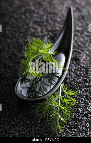 Olio vegetale in una piccola ciotola con aneto rami su uno sfondo di sesamo nero. Foto Stock