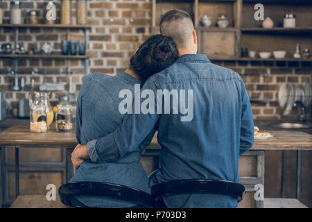 Vista posteriore della coppia giovane in denim shirt abbracciando seduta al tavolo di legno e di avere la prima colazione Foto Stock