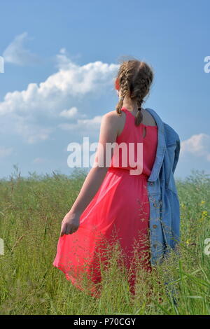 Giovani capelli biondi (breve trecce) ragazza adolescente stand indietro nel prato, vestito in rosa estate abito senza maniche, detiene jeans blu camicia sul suo braccio. Foto Stock