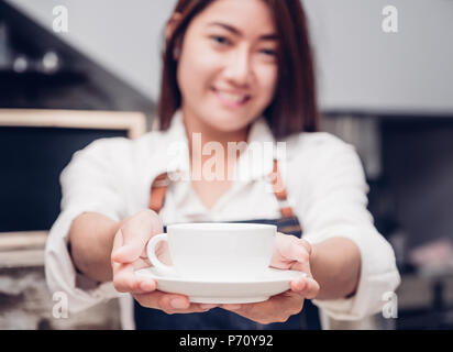 Asia donna barista indossare jean grembiule tenendo calda tazza di caffè servito al cliente con volto sorridente al banco bar,ristorante Cafe concetto di servizio.cameriera Foto Stock