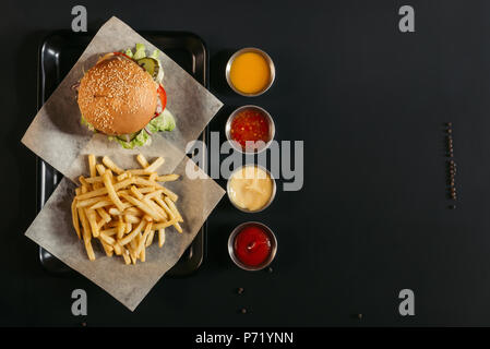Vista dall'alto di patatine fritte con deliziosi burger sul vassoio e salse assortite su nero Foto Stock