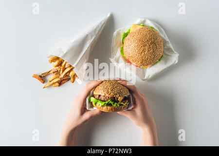 Vista ritagliata della mani con hamburger e patatine fritte nel cono di carta, isolato su bianco Foto Stock