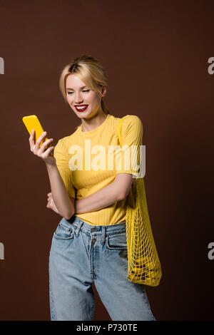 Bella sorridente ragazza bionda con borsa di stringa utilizzando lo smartphone giallo isolato su marrone Foto Stock