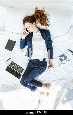 Vista dall'alto di imprenditrice in tuta di parlare al telefono a letto tra cartelle e documenti Foto Stock