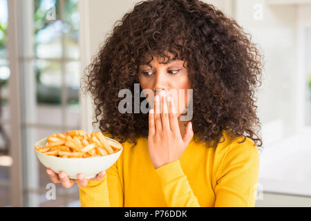 African American donna tenendo una piastra con le patatine a casa coprono la bocca con la mano sconvolto con vergogna per errore, espressione di paura, paura in s Foto Stock