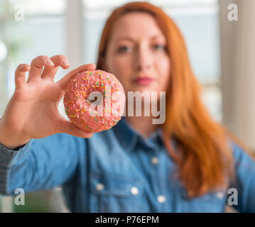 Redhead donna ciambella di contenimento a casa con un fiducioso espressione sul viso intelligente pensare grave Foto Stock