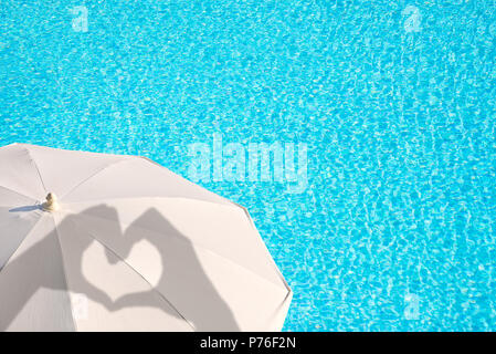 Le ombre delle mani formando un cuore su un ombrellone bianco, blu piscina sullo sfondo dell'acqua, concetto di estate Foto Stock