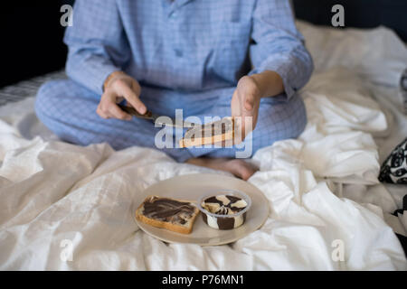 Crop uomo seduto sul letto in pigiama e preparare la colazione diffusione di cioccolato sul pane tostato. Foto Stock