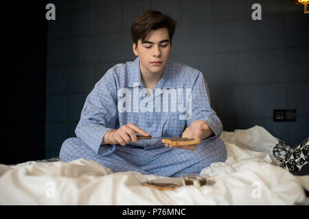Attraente giovane maschio in pigiama seduto sul letto in camera oscura e preparare la prima colazione. Foto Stock