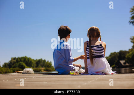 Carino piccoli fratellini seduta sul molo e avere spuntini Foto Stock