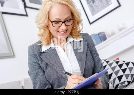 Donna di mezza età psicologo seduta in modo informale home office indossando occhiali la scrittura delle informazioni dei client close-up concentrato gioioso sorridente Foto Stock
