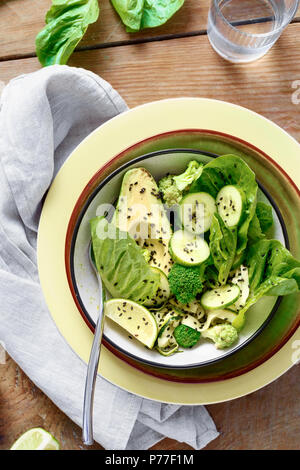 Ciotola estate insalata verde sul tavolo di legno con il vetro di acqua, vista dall'alto. Cibo sano concetto. Insalata di lattuga romana; zucchine, broccoli, avocado, cucum Foto Stock