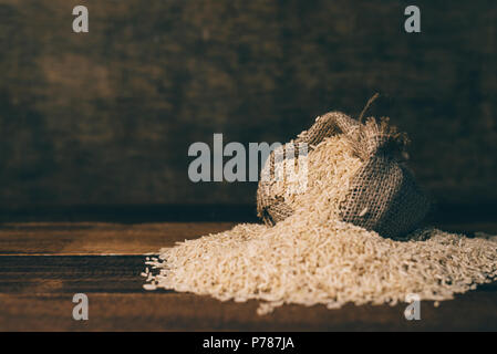Non lucidato naturale riso marrone in un piccolo sacco di tela su tavola in legno su sfondo scuro. cibo sano e mangiare il concetto di messa a fuoco selettiva Foto Stock