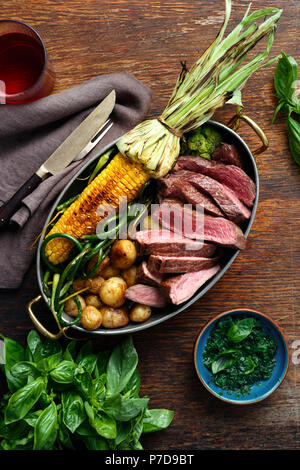 Fette di carne di manzo servita in padella con la frittura di mais, patate e fagioli verdi su sfondo di legno con basilico e salsa di vino rosso, vista dall'alto Foto Stock