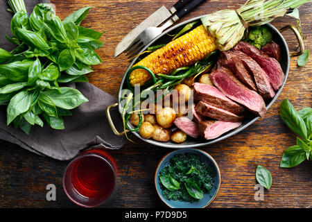 Fette di carne di manzo servita in padella con la frittura di mais, patate e fagioli verdi sul tavolo di legno con salsa di basilico e bicchiere di vino rosso, vista dall'alto Foto Stock