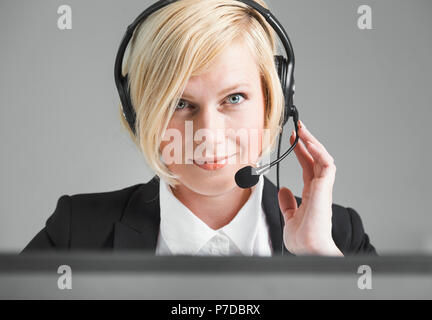 Concentrato di operatore di call center, donna bionda vestita in giacca nera e camicia bianca lavorando sul computer portatile tramite auricolare in carica bianca Foto Stock