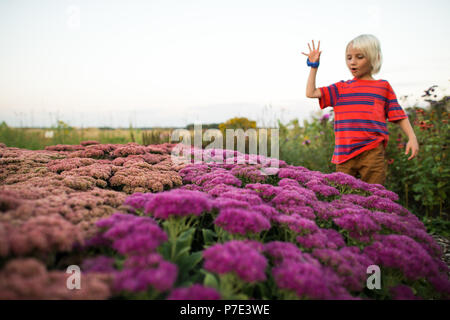 Capelli biondi boy in giardino guardando giù in rosa e fiori viola Foto Stock