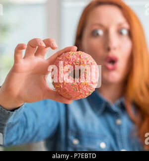 Redhead donna ciambella di contenimento a casa spaventata in stato di shock con una faccia a sorpresa, paura e eccitati con espressione di paura Foto Stock