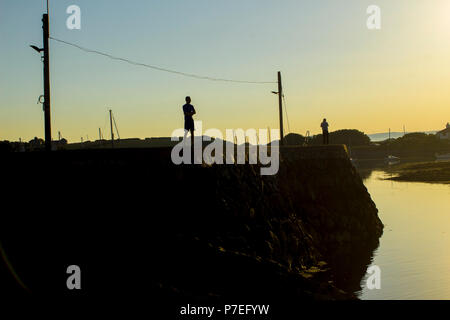 Due uomini in silhouette in piedi su una parete del porto in silhouette contro la calda serata bagliore del sole al tramonto a Groomsport in Irlanda del Nord Foto Stock