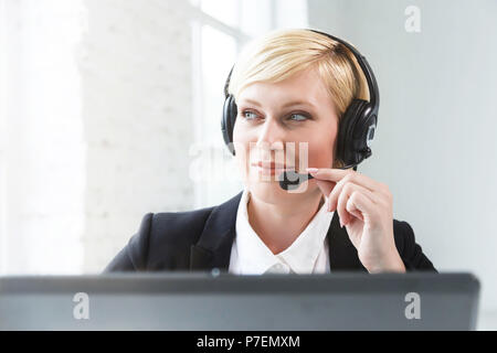 Operatore di call center, donna bionda vestita in giacca nera e camicia bianca lavorando sul computer portatile tramite auricolare in carica bianca Foto Stock